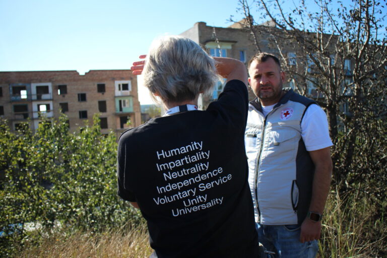 A woman wearing a black t-shirt with the principles of "Humanity, Impartiality, Neutrality, Independence, Voluntary Service, Unity, Universality" printed on the back, shielding their eyes from the sun while speaking with a Red Cross worker in a white vest. The background features damaged buildings and overgrown vegetation.