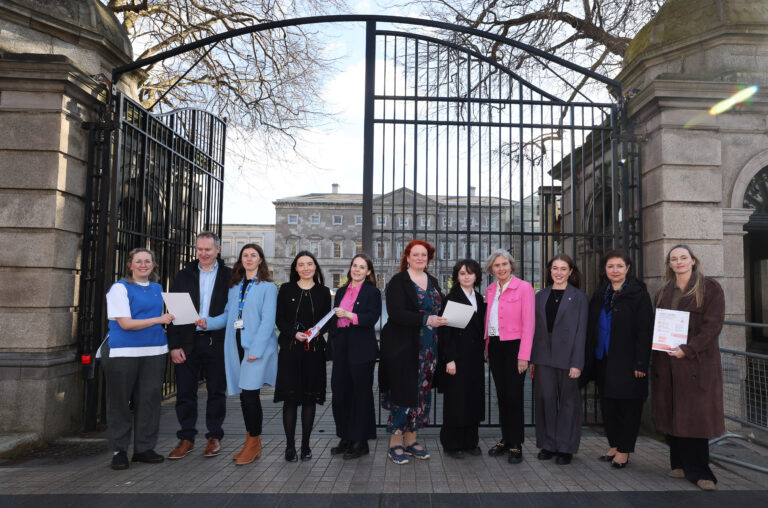 A group of people stand in front of a black iron gate outside a historic stone building, holding documents. They are dressed in professional and semi-formal attire. The sky is clear, and leafless tree branches frame the top of the image.
