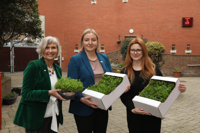 Three women standing outdoors in a courtyard, holding containers of fresh green shamrocks. They are smiling, and a red brick building is visible in the background.